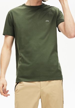 Mies, joka käyttää oliivinvihreää lyhythihasta t-paitaa, jossa on pieni krokotiililogo rinnassa, ja beigejä housuja, säätää hiuksiaan oikealla kädellä.