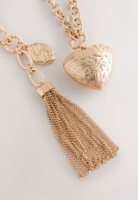 Gold-tone necklace with engraved heart locket and multi-strand chain tassel pendant on a light background.