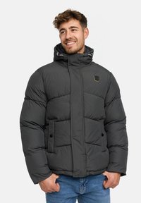 Man die glimlacht, gekleed in een zwarte gewatteerde winterjas met capuchon en blauwe jeans, met zijn handen in zijn zakken, tegen een effen lichte achtergrond.