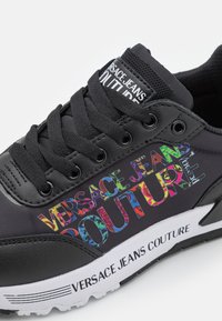 Mustat tennarit, joissa on sekoitus teksturoituja synteettisiä materiaaleja ja monivärinen "Versace Jeans Couture" -logo sivussa sekä valkoinen pohja.