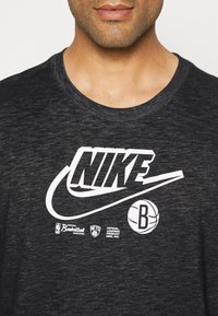 Hombre vistiendo una camiseta de algodón gris oscuro con el logotipo blanco de Nike, los logotipos de la NBA (Asociación Nacional de Baloncesto) y de los Brooklyn Nets, además de un gráfico de baloncesto.