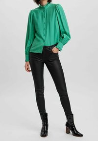 Grüne Bluse mit langen Ärmeln, hohem Kragen und durchgeknöpftem Front. Kombiniert mit schwarzen, enger geschnittenen Hosen und glänzenden Ankle-Boots mit Blockabsatz.