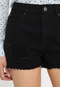 Shorts de mezclilla negra con dobladillos deshilachados, con un parche desgastado en la parte frontal y un diseño clásico de cinco bolsillos.