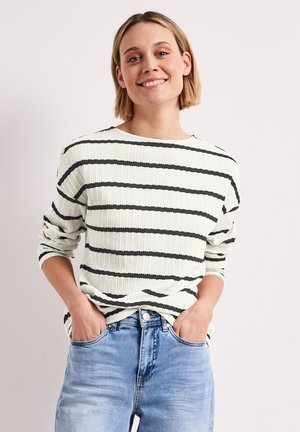 Femme souriante aux cheveux blonds courts portant un pull blanc à rayures horizontales noires et un jean bleu clair, les mains dans les poches.