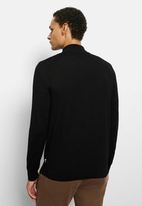 Schwarzer Strickpullover mit langen Ärmeln und hohem Kragen, mit strukturierter Oberfläche und einem kleinen Akzentdetail am Saum.