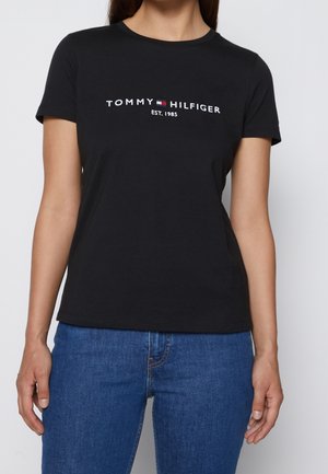 Kvinde iført en sort T-shirt med korte ærmer med "Tommy Hilfiger EST. 1985" logo og blå jeans, stående mod en ensfarvet baggrund.