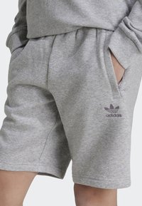 Graue Baumwollshorts mit elastischem Bund, seitlichen Taschen und einem lila Adidas-Logo, das auf dem linken Bein gedruckt ist.