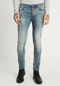 Hombre que lleva puestos unos jeans ajustados de tono azul desgastado con manchas de pintura y un pequeño desgarro en el muslo derecho, combinados con botas negras con cordones y una camiseta de manga larga gris.