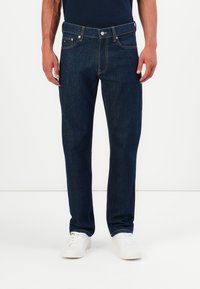 Homme portant un jean droit bleu foncé et des baskets blanches, se tenant devant un fond blanc uni.