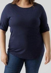 Dunkelblaues, dehnbares Oberteil mit Rundhalsausschnitt und drei Viertelärmeln, an den Bündchen leicht gerafft, aus weichem Material. Kombiniert mit blauen Jeans.