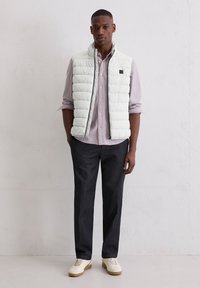 Hellgraue gesteppte Weste mit Stehkragen über einem gestreiften Hemd mit Knöpfen. Dunkle Hose und cremefarbene Sneakers vervollständigen das Outfit.