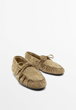 Light brown suede moccasin slippers with bow ties on top, gathered stitching around the toe, and flat black soles.