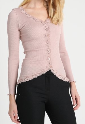 Kvinde iført en tætsiddende støvet lyserød ribbet cardigan med bølget blondestrimle og knapper, kombineret med sorte strukturerede bukser.