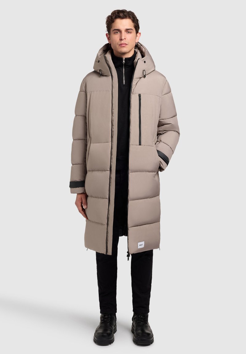 Bēšas krāsas gari piepūsti mantiņu ar kapuci, ar priekšējo rāvējslēdzēju un krūšu kabatu. Melni akcenti uz piedurknēm un logotipa patch tuvumā apakšmalai.