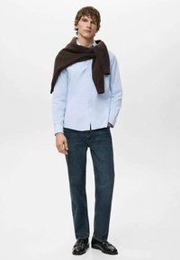 Jasnoniebieska koszula zapinana na guziki z ciemnobrązowym swetrem zarzuconym na ramiona, w połączeniu z ciemnoniebieskimi dżinsami i czarnymi butami.