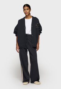 Großer schwarzer Pullover mit Knöpfen und Taschen, kombiniert mit weiten schwarzen Hosen mit seitlichen Streifen. Einfaches weißes T-Shirt darunter.