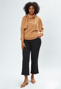 Beige Pullover mit drapiertem Ausschnitt und Verzierungen, kombiniert mit schwarzen Cropped-Hosen und braunen Mules mit dekorativen Akzenten.