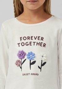 Witte langhangende shirt met lange mouwen, versierd met geborduurde bloemen in paars, blauw en wit, met de tekst "VOOR ALTIJD SAMEN" en "SASSY SQUAD" eronder.