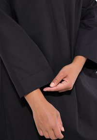 Sort oversized kjole med lange ærmer. Nærbillede viser rullemanchet, glat tekstur, og en hånd der justerer stoffet ved ærmet.