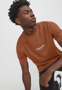 Camiseta de algodón marrón con cuello redondo, que presenta un logo blanco de "Originals Studio" en el pecho, combinada con jeans oscuros y zapatillas.