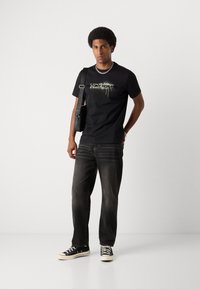 Tricou negru cu "ANTONY MORATO" imprimat într-un design cu picături, asociat cu blugi negri decolorati și adidași negri. Lănțișor argintiu și geantă.