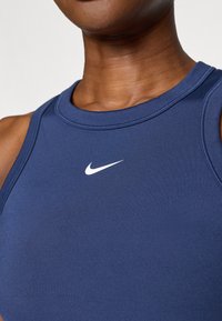 Námornícky modrý športový top bez rukávov vyrobený z hladkého materiálu, s bielym logom Nike na prednej strane a okrúhlym výstrihom.