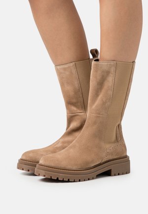 Bottes chelsea en daim marron clair avec des panneaux élastiques sur les côtés et une semelle en caoutchouc texturée. Équipées d'une languette pour un enfilage facile et de perforations décoratives.