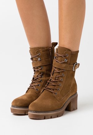 Bottines en daim marron à lacets avec fermeture éclair latérale, sangle, boucle et talon épais portées sur des jambes nues sur fond blanc.
