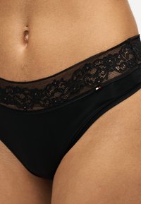 Gros plan d'une personne portant des sous-vêtements noirs avec une ceinture en dentelle florale et un petit logo rectangulaire sur le côté.