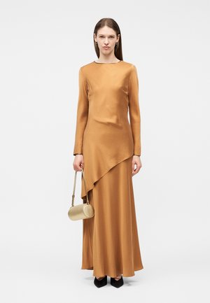 Γυναίκα με μακρύ σατέν φόρεμα σε χρυσοκαφέ απόχρωση με ασύμμετρο τελείωμα, κρατώντας μια μικρή χρυσή κυλινδρική τσάντα, στέκεται μπροστά σε λευκό φόντο.