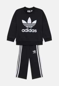 Μαύρη αθλητική μπλούζα Adidas με λευκό λογότυπο και τρεις ρίγες στα μανίκια, συνδυασμένη με μαύρο παντελόνι που διαθέτει λευκές ρίγες και λογότυπο.