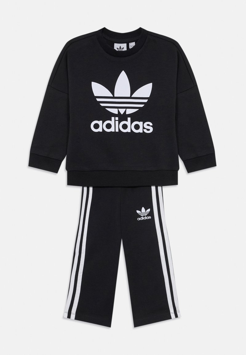 Melna Adidas džemperis ar baltu logo un trim svītrām uz piedurknēm, saskaņots ar melniem biksēm, kas rotātas ar baltām svītrām un logo.