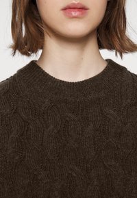 Dunkelbrauner gestrickter Pullover mit einem strukturierten Wellenmuster und einem gerippten Ausschnitt, der aus einem weichen, gemütlichen Material gefertigt ist.