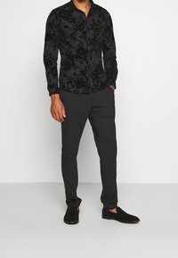 Zwarte blouse met bloemenpatroon, gemaakt van gestructureerde stof, gecombineerd met donkere broek en zwarte schoenen, wat zorgt voor een gecoördineerde monochrome look.