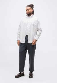 Άνδρας με λοβούς μαλλιών που φοράει λευκό πουκάμισο με ρίγες και κουμπιά πάνω από λευκή μπλούζα, σκούρο γκρι παντελόνι, λευκές κάλτσες και σκούρα καφέ μοκασίνια, όρθιος.