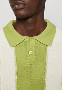 Gehaakt poloshirt met een groen en crèmekleurig dambordpatroon, voorzien van een gestructureerde kraag en twee groene knopen bij de sluiting.