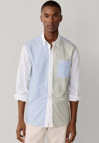 Uomo che indossa una camicia a maniche lunghe abbottonata con strisce verticali blu e beige e maniche bianche arrotolate, in piedi davanti a uno sfondo semplice.