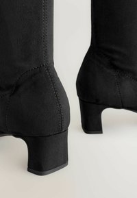 Bottines noires en matière suédée, avec un talon épais et une texture lisse, présentant des détails de couture visibles.