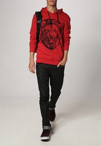 Roter Kapuzenpullover mit schwarzem Löwengrafik, gerippten Bündchen und Saum. Kombiniert mit eng anliegenden schwarzen Hosen und burgunderfarbenen Sneakers.
