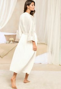 Longue robe blanche avec des accents en dentelle sur les manches, une taille ceinturée, une jupe fluide et des manches retroussées ; texture douce sur un fond neutre.