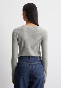 Vrouw met lang, donker haar die een lichtgrijze geribde lange mouwtop en hoge taille, donkerblauwe spijkerbroek draagt, gezien van achteren.