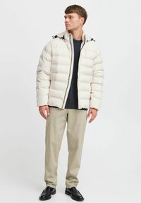 Puffjakke i lys beige, med hette, glidelås og subtilt quiltet design. Kombinert med beige bukser og svarte sko.