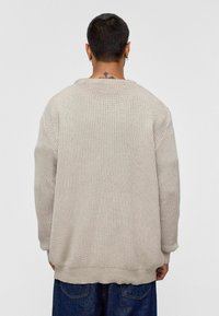 Beige gebreide trui met een losse pasvorm, ribbelstructuur en verlaagde schouders, gecombineerd met donkere spijkerbroek. Voorzien van een ronde halslijn.