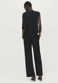 Vrouw staand met haar rug naar voren, gekleed in een zwart mouwloos topje met geplooide details, wijde broek, zwarte hakken en een gouden armband.