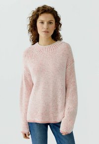 Junge Frau mit lockigem Haar, die einen rosa-weiß gestreiften Pullover und blaue Jeans trägt, steht vor einem einfarbigen hellen Hintergrund.