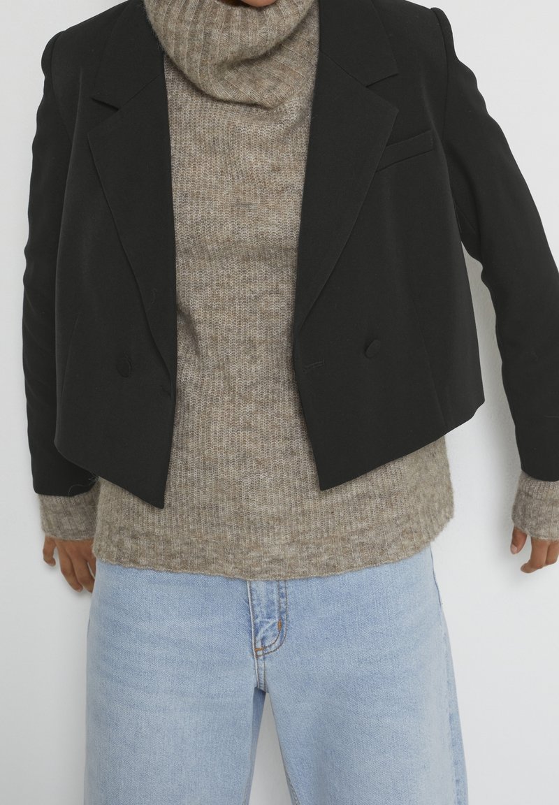 Musta lyhyt bleiseri, jossa on kaksirivinen napp closure, yhdistetty vaalean beige ribbed poolopaitaan ja vaaleansiniisiin farkkuihin. Teksturoitu kangas näkyvissä.