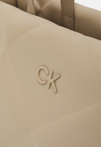 Kreminės spalvos dirbtinės odos rankinė su siuvinėta dizainu ir iškilniu "CK" logo. Lygus paviršius su dviem rankenom nešimui.