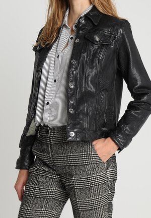 Mujer que lleva una chaqueta de cuero negra sobre una camisa blanca y negra de rayas, combinada con pantalones de cuadros en blanco y negro, mano en el bolsillo.