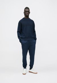 Tamsiai mėlynas megztinis su apvalia iškirpte, derinamas su prigludusiomis siaurėjančiomis kelnėmis ir baltais sportbačiais su rudais padais. Paprastas, minimalistinis dizainas.