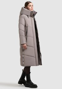 Lys grå pufferjakke med høj krave, der har kviltede sektioner, lynlåse og trykknapper langs siden. Stylet med sorte ankelstøvler.